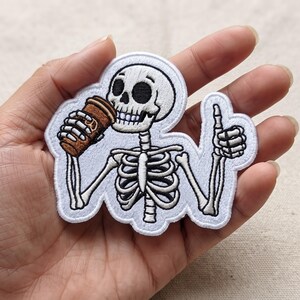 May include: An embroidered patch featuring a white skeleton with black outlines. The skeleton holds a brown coffee cup to its mouth with one hand and gives a thumbs-up gesture with the other hand. The design is set against a white background.