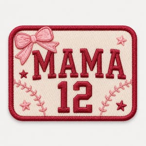May include: A rectangular embroidered patch with a cream-colored background and a dark red border. The patch features the text MAMA 12 in dark red, a pink embroidered bow in the top left corner, and small pink stars and baseball stitching accents.