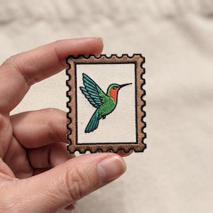 May include: A rectangular embroidered patch shaped like a postage stamp with a scalloped edge. The patch features a central design of a hummingbird with green wings, a red throat, and a black beak, set against a cream-colored background.