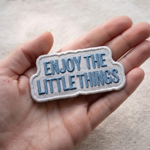 May include: A rectangular embroidered patch with a light beige fabric base and a white stitched border. The text "ENJOY THE LITTLE THINGS" is embroidered in a bold, medium blue thread across two lines in a clean, sans-serif font style.