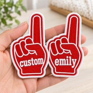 May include: Two red and white embroidered patches shaped like foam fingers. The left patch features the word custom in white lowercase letters, and the right patch features the word emily in white lowercase letters, both with a white border.