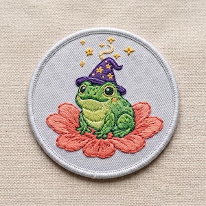 May include: A circular embroidered patch on a light gray background featuring a green frog wearing a purple wizard hat with yellow stars. The frog sits atop a large coral-colored flower, surrounded by small yellow embroidered stars above the hat.