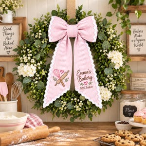 May include: A round green wreath featuring a large pink and white striped fabric bow with scalloped edges. The bow tails display a pink whisk, a wooden rolling pin, and the text Emma's Bakery best baker with small pink heart accents.