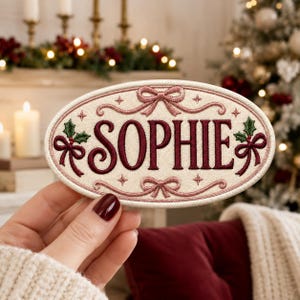 May include: An oval-shaped embroidered patch with a cream-colored background featuring the name SOPHIE in dark red capital letters. The design includes decorative pink bows, swirling lines, small star accents, and green holly leaves with red berries.
