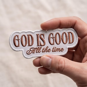May include: A white embroidered patch with a scalloped edge featuring the brown text "GOD IS GOOD" in a bold, sans-serif font on the top line and the brown cursive script "All the time" on the bottom line, creating a rectangular shape.