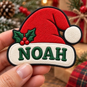 May include: A red and white embroidered patch shaped like a Santa hat with a white pom-pom. The white brim features the name NOAH stitched in dark green thread. Two green holly leaves with three red berries are attached to the side of the hat.