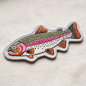May include: An embroidered iron-on patch shaped like a rainbow trout. The fish features an olive green back with dark spots, a vibrant pink lateral stripe, a white underbelly, and orange fins. The patch has a light gray border outline.