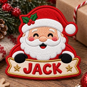 May include: An embroidered patch featuring a smiling Santa Claus wearing a red hat with white trim and a holly sprig. Santa has a white beard, pink cheeks, and black mittens holding a beige banner with the name JACK in red and two gold stars.