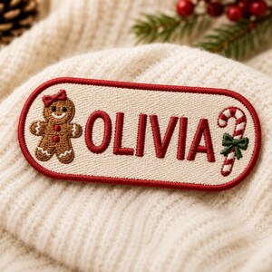 May include: An oval-shaped embroidered patch with a red border on a cream background. The patch features the name OLIVIA in red capital letters, flanked by a brown gingerbread person with a red bow and a red and white candy cane with a green bow.