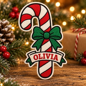 May include: An embroidered candy cane patch featuring diagonal red and white stripes, a green bow tied around the center, and a white banner at the bottom with the name OLIVIA stitched in red capital letters with a green border.