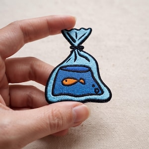May include: An embroidered patch featuring a light blue plastic bag tied at the top, containing a dark blue fishbowl with a small orange goldfish swimming inside. The design is outlined in black thread with visible stitching details throughout.