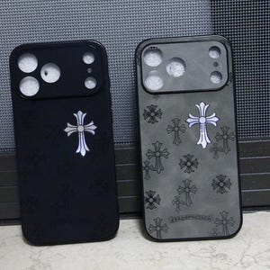 May include: Two rectangular smartphone cases with camera cutouts. The left case is solid black with a single silver cross emblem. The right case is grey with a repeating pattern of black and silver cross symbols and a small logo at the bottom.