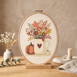 Puede incluir: Un bastidor de bordado ovalado de madera que contiene un patrón de punto de cruz de una taza color crema con un corazón rojo oscuro, llena de hojas y flores de otoño, flanqueada por una calabaza naranja y una pequeña calabaza blanca.