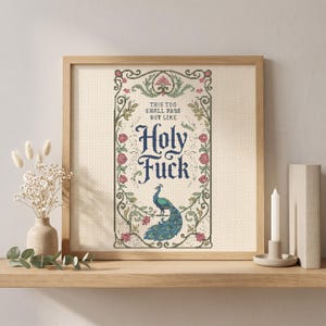 May include: A framed cross-stitch embroidery featuring a decorative border of green vines and pink roses surrounding a peacock illustration. The text reads: This too shall pass but like Holy Fuck. The design is centered on a light-colored fabric.
