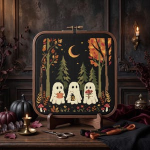 May include: A square cross-stitch embroidery in a wooden hoop featuring three white ghosts in a dark forest at night. The scene includes a yellow crescent moon, evergreen trees, autumn foliage, red mushrooms, and ghosts holding a pumpkin, lantern, and flowers.