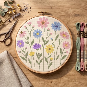May include: A circular wooden embroidery hoop containing a cross-stitch pattern of various colorful wildflowers in shades of pink, blue, yellow, and purple on a cream-colored fabric, accompanied by metal scissors and three skeins of embroidery floss.