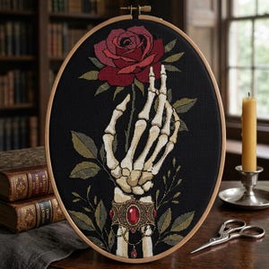 Könnte beinhalten: Ein ovaler Stickrahmen mit einem Kreuzstichmuster einer skelettierten Hand, die eine verzierte goldene Manschette mit einem großen roten Edelstein trägt. Die Hand hält eine einzelne dunkelrote Rose mit grünen Blättern auf schwarzem Stoff.