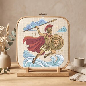 May include: A square embroidery hoop containing a cross-stitch design of a warrior in golden armor with a red cape, holding a spear and a round shield featuring a lion emblem, leaping over stylized blue waves against a cream-colored fabric background.