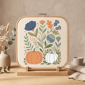 Puede incluir: Un bordado de punto de cruz cuadrado en un marco de madera sobre soporte, con un diseño botánico que incluye una calabaza naranja grande, una calabaza blanca, flores azules, hojas naranjas y follaje verde sobre un fondo de tela color crema.
