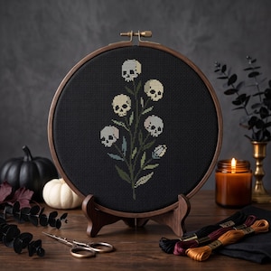 May include: A circular wooden embroidery hoop containing black fabric with a cross-stitch design of a plant with off-white skull-shaped flowers and green leaves. The hoop is mounted on a small wooden stand, displaying a detailed needlework piece.