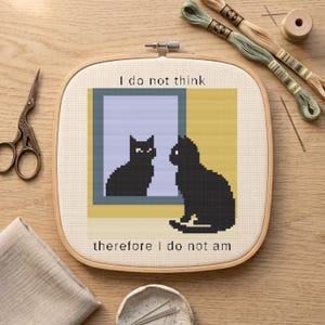 May include: A square embroidery hoop containing a cross-stitch pattern of a black cat sitting before a window, with its reflection visible in the glass. The text "I do not think" appears above the window and "therefore I do not am" below.