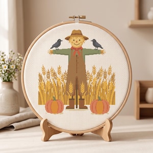 May include: A circular wooden embroidery hoop containing a cross-stitch pattern of a scarecrow wearing a hat and red scarf, standing in a field of golden wheat with two black crows perched on its arms and two orange pumpkins at its base.
