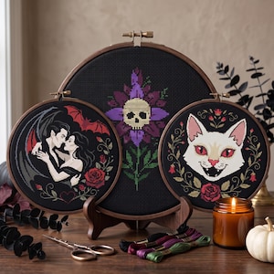 May include: Three circular wooden embroidery hoops containing cross-stitch designs on black fabric. One hoop features a vampire couple with a bat, another a skull centered on a purple flower, and the third a white cat face surrounded by red roses.