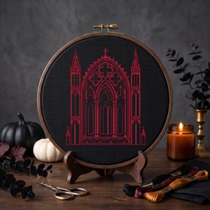 May include: A circular wooden embroidery hoop containing black fabric with a cross-stitched design of a red gothic cathedral window. The hoop is mounted on a small wooden stand, showcasing intricate architectural details in the needlework.