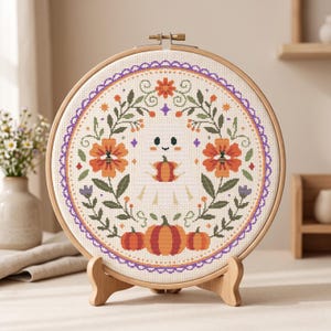 Puede incluir: Un bastidor de bordado circular de madera con un patrón de punto de cruz de un pequeño fantasma blanco sosteniendo una calabaza. El diseño presenta una corona floral con flores naranjas, hojas verdes y tres calabazas en la base, con un borde morado.