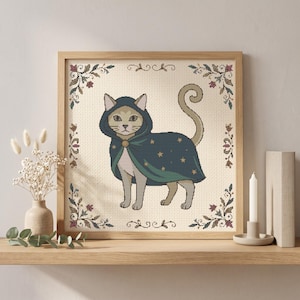 May include: A framed cross-stitch embroidery featuring a light-coloured cat wearing a dark teal hooded cloak decorated with gold stars. The cat is centred on a cream-coloured fabric background, framed by delicate floral corner motifs in muted tones.