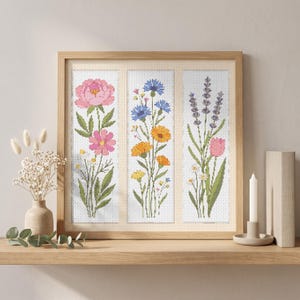 May include: A framed cross-stitch embroidery featuring three vertical panels of botanical illustrations. The left panel shows pink peonies and cosmos, the center displays blue cornflowers and orange marigolds, and the right depicts lavender and a pink tulip.