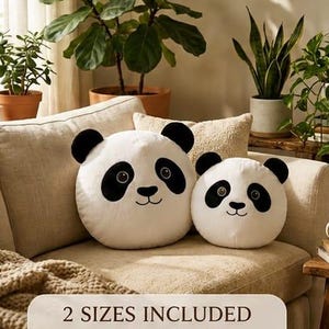 Op de afbeelding: Twee ronde, panda-vormige pluche kussens in wit en zwart. De kussens zijn voorzien van geborduurde gezichtsdetails, waaronder ogen, een neus en een mond. Een rechthoekig label onderaan toont de tekst: 2 SIZES INCLUDED BIG & SMALL.