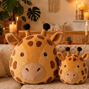 May include: Two round, plush giraffe toys in varying sizes, featuring a golden-yellow fabric with brown spots, dark brown ossicones, and embroidered facial features including round eyes and a snout, positioned side by side on a patterned sofa.