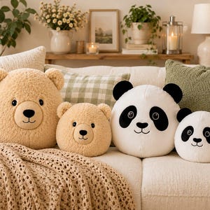 Könnte beinhalten: Ein Set aus vier runden Plüschkissen, bestehend aus zwei beigefarbenen Bären mit flauschiger Textur und zwei weißen Pandas mit schwarzen Ohren und Augenpartien. Jedes Kissen hat gestickte Gesichtsmerkmale wie Augen, Nase und Mund.