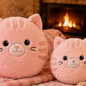 Könnte beinhalten: Zwei runde, hellrosa Plüsch-Katzenkissen mit strukturiertem, flauschigem Stoff. Jedes Kissen verfügt über gestickte Gesichtsdetails, darunter schwarze Augen, eine kleine rosa Nase, einen gestickten Mund, Schnurrhaare und drei Streifen.