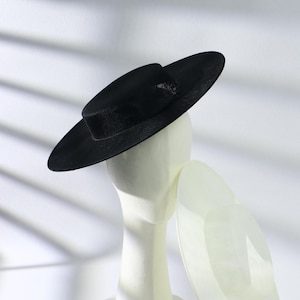 May include: A black boater hat with a flat crown and a wide, stiff brim. The hat is constructed from a woven material with a visible texture and is displayed on a white mannequin head, showcasing its structured shape and clean, circular silhouette.