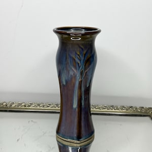 May include: A tall, slender ceramic vase with a flared rim, featuring a glossy glaze that transitions from a deep, dark brown at the base to a vibrant, mottled blue and purple pattern that drips downward along the body of the vessel.