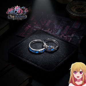 May include: Two silver-toned rings featuring star-shaped accents and blue glowing details. One ring has a smooth band with blue star inlays, while the other features a cluster of small clear stones and blue stars. The text Oshi no Ko 3rd Season is visible.