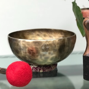 May include: A bronze-colored metal singing bowl with a hammered texture and a circular etched symbol on the side. The bowl rests on a patterned fabric cushion and is accompanied by a wooden mallet with a bright red felt ball and a wooden striker.