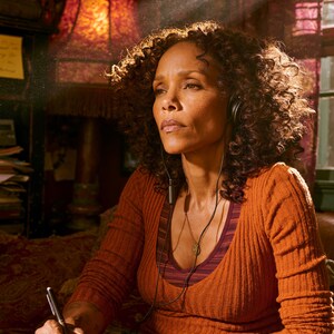May include: A person with curly hair wears a ribbed orange long-sleeve shirt and a dark striped tank top. They have black wired headphones around their neck, a pendant necklace, and stacked bracelets. They are writing in a yellow notebook with a pen.