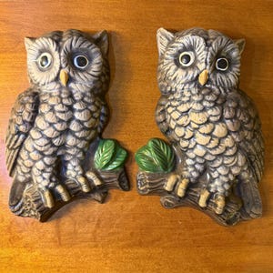 May include: Two wall plaques shaped like owls perched on tree branches. Each owl features textured grey and brown feathers, large round eyes with black pupils, and a yellow beak. A small cluster of green leaves is attached to each branch.