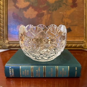 May include: A clear, cut-glass bowl featuring a scalloped rim and a geometric diamond pattern. The bowl is resting on a dark green Reader's Digest book titled The Third Twin, Small Town Girl, To The Hilt, and The Running Man, Volume 2 1997.