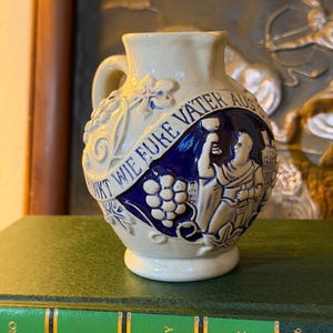 May include: A beige ceramic pitcher featuring a blue relief panel with a figure holding a goblet and a cluster of grapes. The pitcher has a raised banner with the German text WIE EUKE VATER AUS, surrounded by embossed leaf and vine patterns.