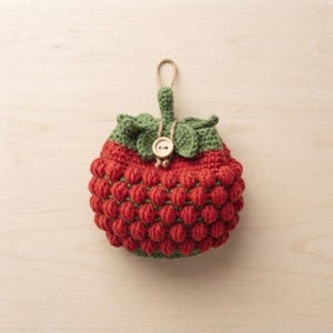 Puede incluir: Un bolso de ganchillo redondo con forma de fresa, que presenta un cuerpo rojo texturizado hecho con puntos de burbuja, una parte superior de hojas verdes, un pequeño lazo para colgar y un botón de madera claro centrado en el cierre frontal.