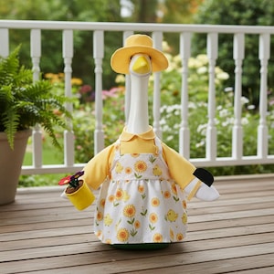 Könnte beinhalten: Eine weiße Gänsefigur mit einem gelben Sonnenhut, einem passenden gelben Langarmshirt und einer weißen Schürze mit einem Muster aus gelben Küken und orangefarbenen Sonnenblumen. Die Gans hält einen kleinen gelben Topf mit Filzblumen.
