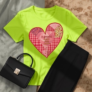 Op de afbeelding: Een neongeel t-shirt met korte mouwen en een groot hartmotief bestaande uit diverse rood en roze patronen. De tekst "A grateful heart sees more" staat in het midden van het hart gedrukt. Een zwarte handtas is eronder geplaatst.