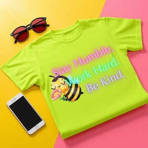 Peut inclure: Un t-shirt à manches courtes vert citron néon avec une abeille dessinée tenant une fleur rose et le texte "Stay Humble. Work Hard. Be Kind." en dégradé. Des lunettes de soleil noires à verres rouges et un smartphone sont placés à côté.