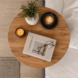 Pu&ograve; includere: Un tavolino rotondo in legno con finitura a venature color marrone chiaro. Sul tavolo si trovano un vaso in ceramica bianca con una pianta verde, una tazzina da caff&egrave; nera con latte art su un piattino, una candela in vetro ambrato accesa e un libro.