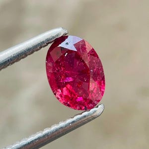 Puede incluir: Una gema de corte ovalado con un color rojo intenso y una superficie facetada, sostenida por unas pinzas de metal plateado. La piedra muestra reflejos de luz internos y bordes definidos en toda su forma pulida y simétrica.