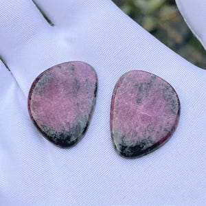 May include: Two polished, teardrop-shaped rhodonite cabochons featuring a smooth surface with a distinct color pattern of mottled pink and deep black mineral inclusions. The stones have a rounded, asymmetrical shape and a glossy, reflective finish.